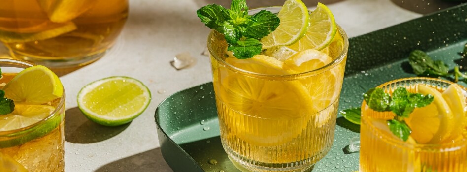 Three cups of iced tea on a green tray, each with lime and lemon slices and herb garnish.  Next to it is a slice of lime and a pitcher of iced tea. 
